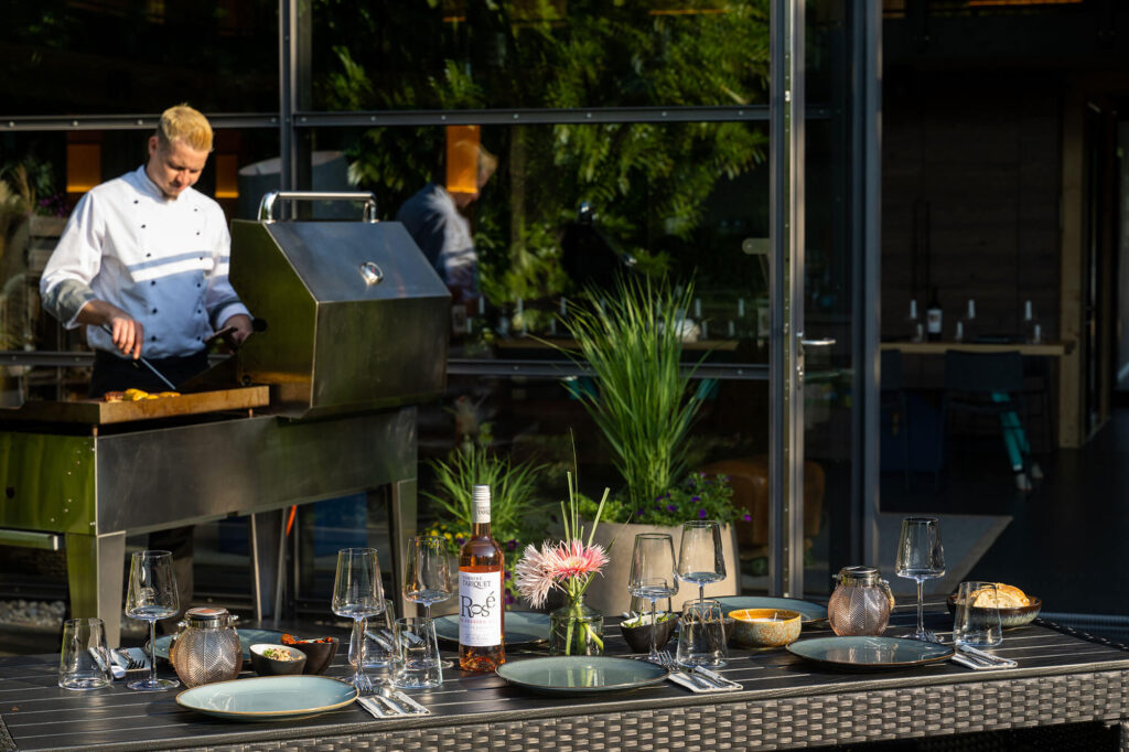 Koch grillt Fleisch an Grillstation, im Vordergrund gedeckter Tisch mit Roséwein, Gläsern, Brot und Blumen.