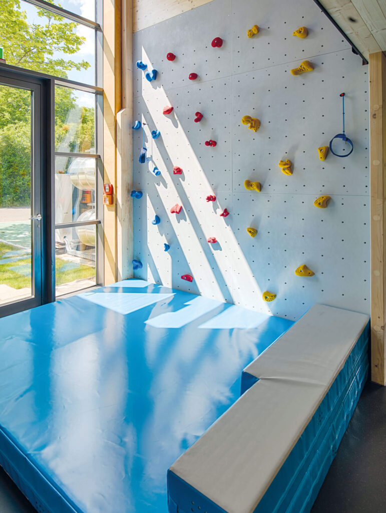 Indoor-Kletterwand mit bunten Griffen und weicher blauer Matte, beleuchtet durch Tageslicht aus großen Fenstern.