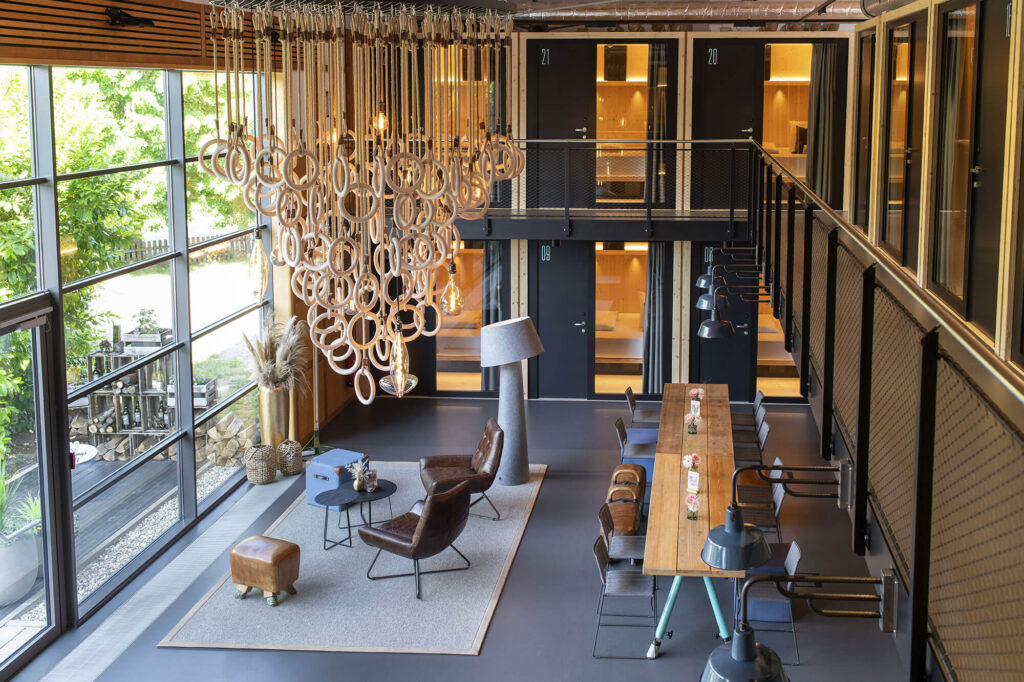 Moderner Lounge- und Essbereich mit Blick in die Schlafpods, großer Fensterfront und auffälligem Holzring-Leuchtobjekt.