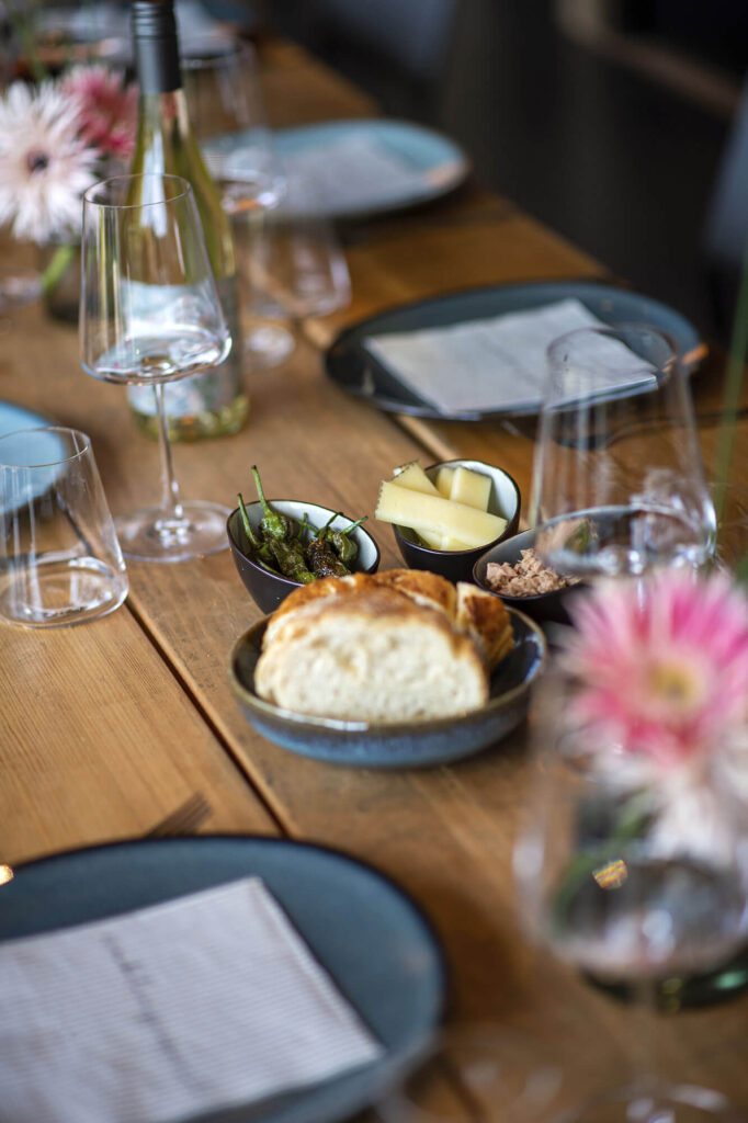 Nahaufnahme eines gedeckten Holztisches mit Brot, Käse, Tapas, Weingläsern und Blumen in gemütlicher Atmosphäre.