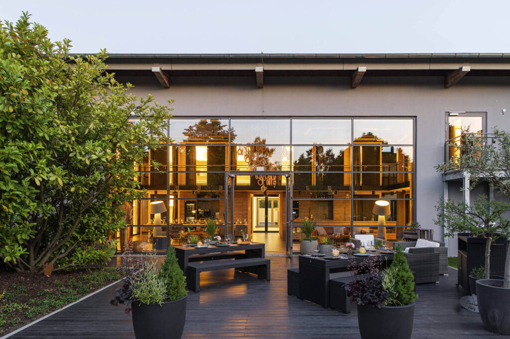 Große Terrasse mit Sitzgruppen vor moderner Glasfassade, dahinter gemütlich beleuchteter Innenraum mit Kronleuchter.