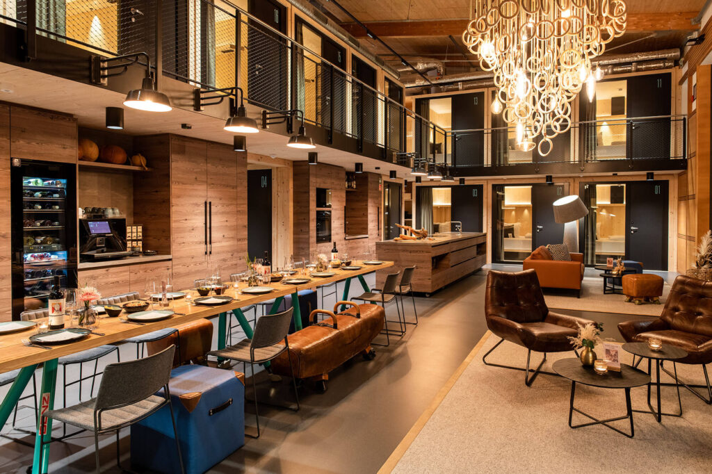 Modern gestalteter Gemeinschaftsraum mit langer Tafel, Lounge-Ecke, Küche und Zugang zu Zimmern auf zwei Etagen.