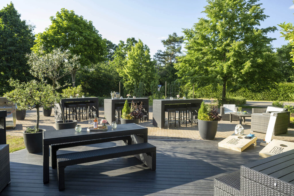Große Terrasse mit modernen Sitzgruppen, Pflanzen und Spielbereich, umgeben von Bäumen bei sonnigem Wetter.
