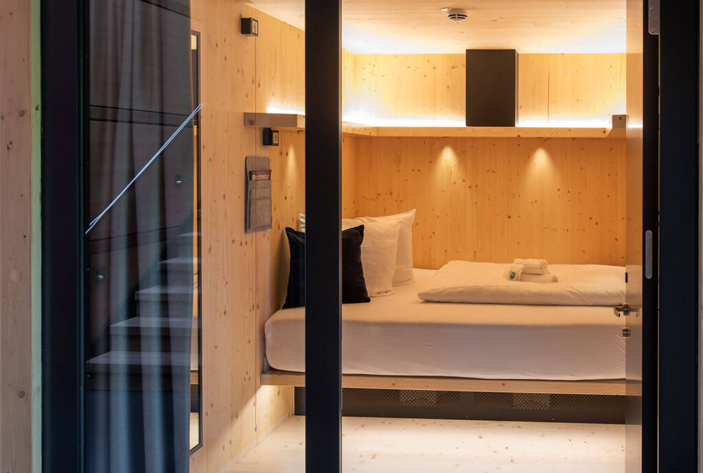 Modernes, gemütlich beleuchtetes Zimmer mit Holzwänden, Einzelbett, Kissen und Handtüchern, Blick durch Glastür.