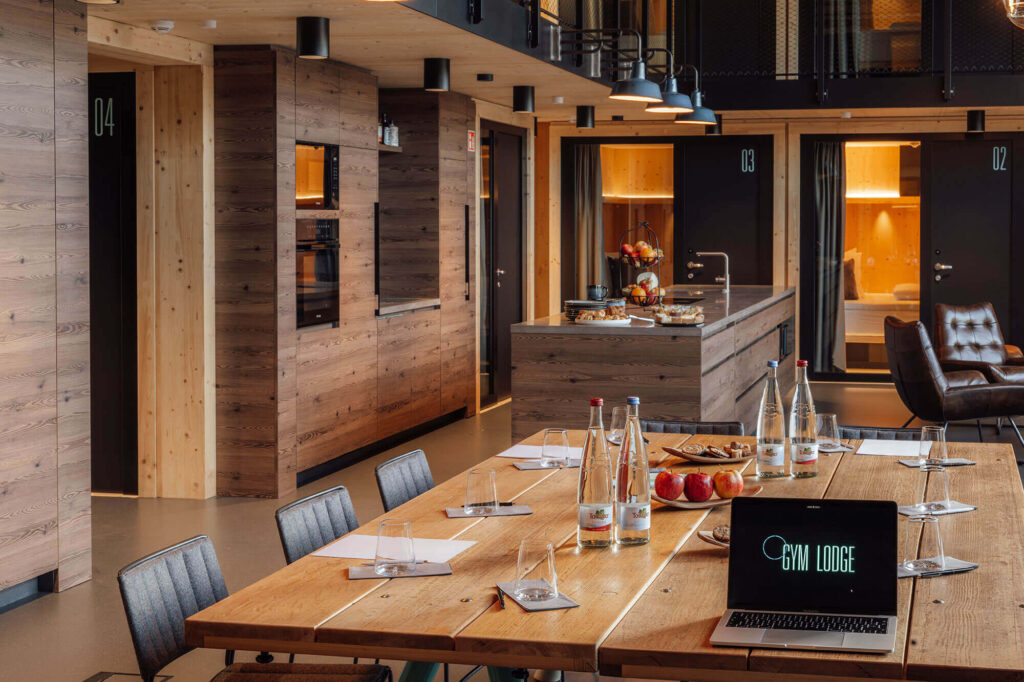 Moderne Gemeinschaftsküche mit Holzelementen, großer Esstisch mit Getränken, Obst und Laptop mit „Gym Lodge“-Logo.