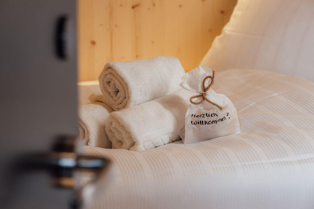Gerollte Handtücher und ein Willkommensgeschenk mit der Aufschrift „Herzlich Willkommen“ auf einem gemachten Bett.