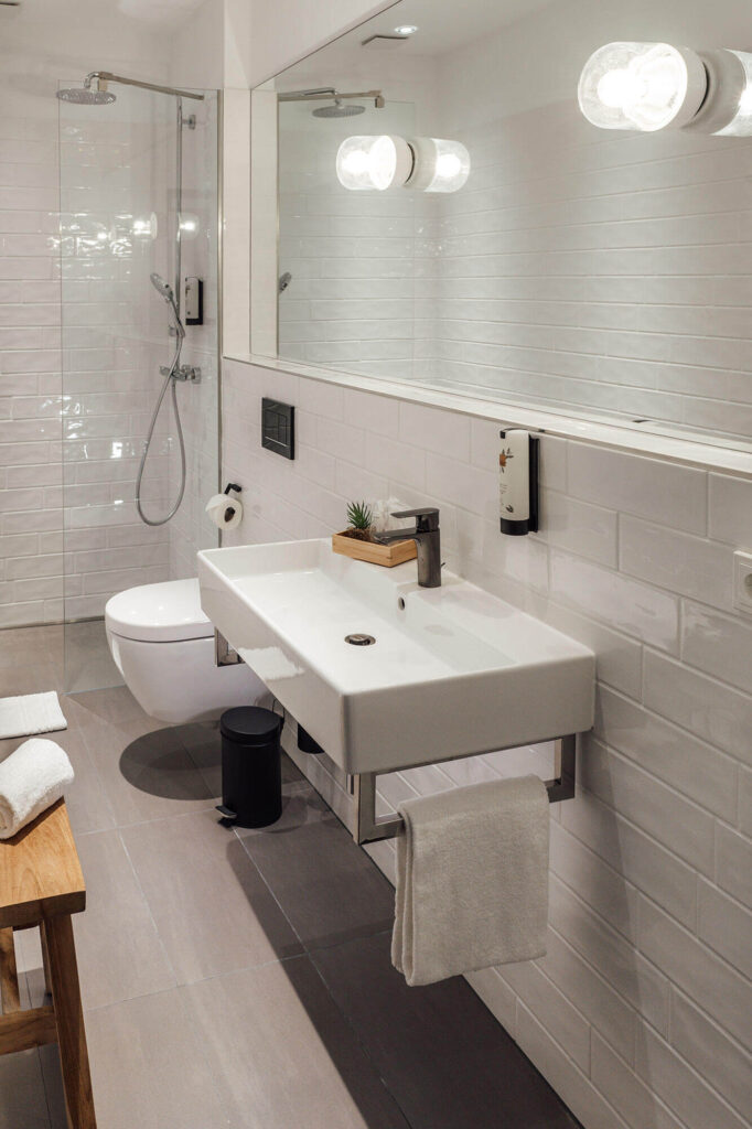 Modernes Bad mit Dusche, WC und Waschbecken vor weiß gefliesten Wänden, Spiegel und Seifenspender an der Wand.