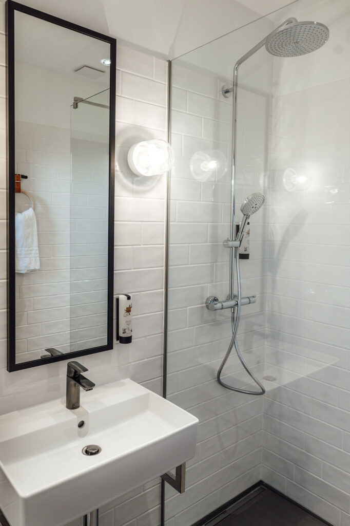 Modernes Bad mit weiß gefliester Dusche, Regendusche, Waschbecken, Spiegel und Wandspender für Seife oder Shampoo.