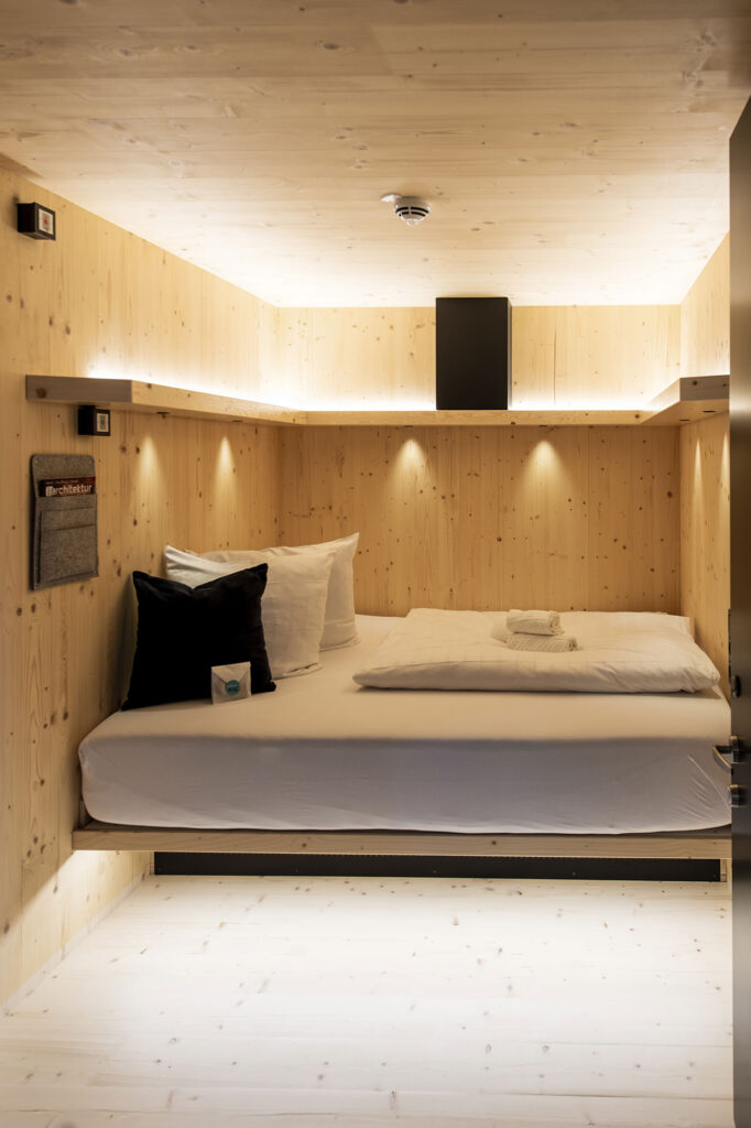 Gemütliche Schlafnische aus hellem Holz mit Doppelbett, Kissen, Handtüchern und indirekter Beleuchtung.